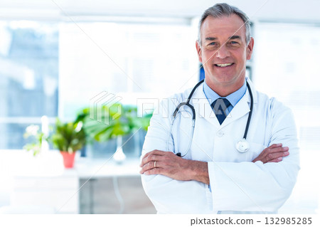 Mature adult male doctor standing arms crossed in coat by windows holding stethoscope, copy space 132985285