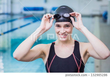 Woman standing at poolside in center pool wearing black swim cap and one-piece suit holding goggles Woman standing at poolside in center pool wearing black swim cap and one-piece suit holding goggles 132985286