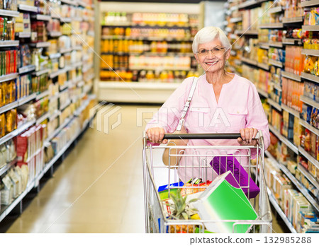 Senior woman pushing shopping cart filled with pineapple and cereal boxes through supermarket aisle 132985288