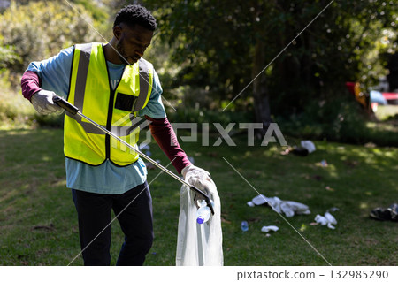 African American man picking up litter in park with metal picker and plastic-lined bag, copy space 132985290