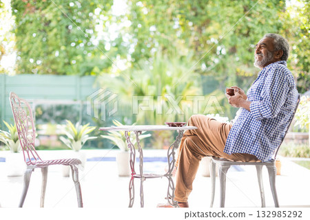Senior African American man sitting at metal bistro table on backyard patio holding cup, copy space 132985292