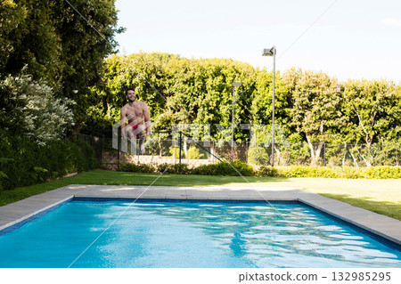 Man jumping from grass lawn into rectangular pool wearing red trunks in backyard garden, copy space Man jumping from grass lawn into rectangular pool wearing red trunks in backyard garden, copy space 132985295