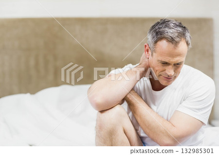 Middle-aged man sitting on bed pressing hand to neck wearing pajama pants with white pillows 132985301