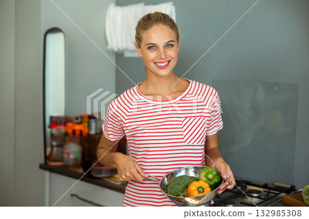 Woman holding metal colander filled with broccoli and bell peppers in kitchen near spice jars Woman holding metal colander filled with broccoli and bell peppers in kitchen near spice jars 132985308