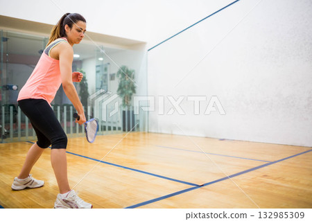 Woman swinging racket while aiming at squash ball on squash court with glass wall background 132985309