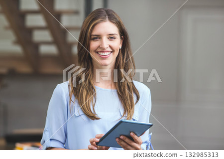 Woman standing in office wearing blue blouse holding tablet near wooden staircase and panel door 132985313