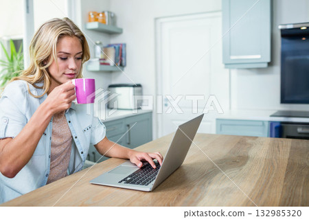 Woman typing on laptop while holding pink mug in modern kitchen with toaster and glass jars 132985320