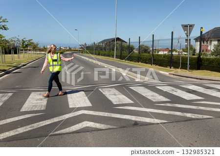 Woman signaling traffic while wearing safety vest and crossing street at zebra crossing 132985321