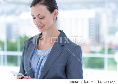 Businesswoman checking smartphone while standing at office windows overlooking city, copy space Businesswoman checking smartphone while standing at office windows overlooking city, copy space 132985322