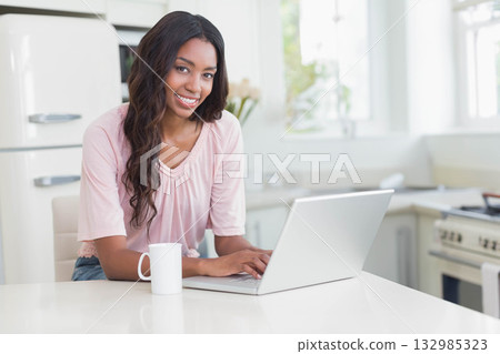 African American woman typing on silver laptop at white kitchen island near ceramic mug and flowers African American woman typing on silver laptop at white kitchen island near ceramic mug and flowers 132985323