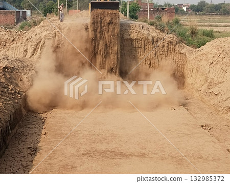 JCB machine dumping loose soil into trench on rural road, creating heavy dust cloud during road construction work in Indian village infrastructure development scene 132985372