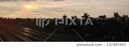 Cloudy evening sky with sun setting over railway lines and silhouetted buildings in the distance 132985403
