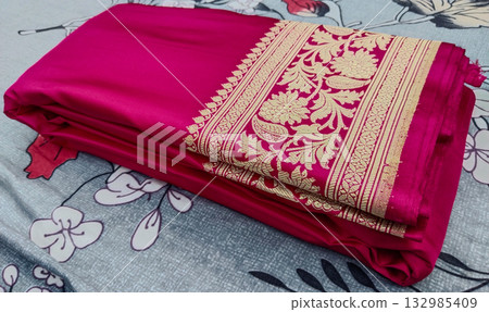 Folded dark pink Banarasi silk saree with intricate golden zari floral border placed on a floral bedsheet, showcasing rich traditional Indian textile and ethnic wedding fashion elegance Folded dark pink Banarasi silk saree with intricate golden zari floral border placed on a floral bedsheet, showcasing rich traditional Indian textile and ethnic wedding fashion elegance 132985409