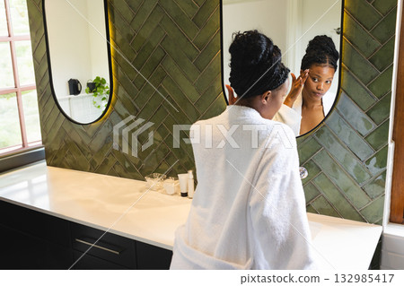 African American woman in white bathrobe applying facial serum at bathroom countertop with mirrors African American woman in white bathrobe applying facial serum at bathroom countertop with mirrors 132985417