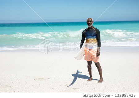 African American senior man standing on beach wearing swim shirt holding surfboard, copy space 132985422