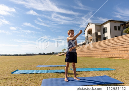 Boy stretching arms on three blue exercise mats on grassy field with wooden bleachers, copy space Boy stretching arms on three blue exercise mats on grassy field with wooden bleachers, copy space 132985432