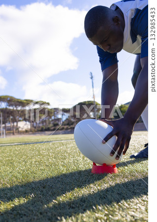 African American male athlete in jersey positioning rugby ball on kicking tee on pitch, copy space 132985433