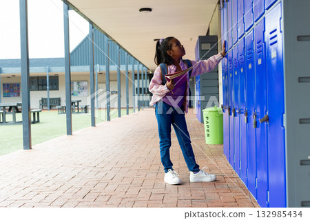 African American schoolgirl opening blue locker, holding notebook on school walkway, copy space 132985434
