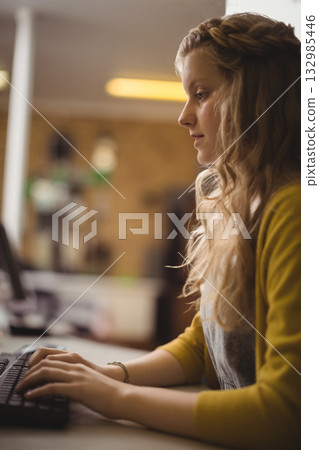 Female child wearing yellow cardigan and bracelet typing on keyboard at desk near bookshelves Female child wearing yellow cardigan and bracelet typing on keyboard at desk near bookshelves 132985446