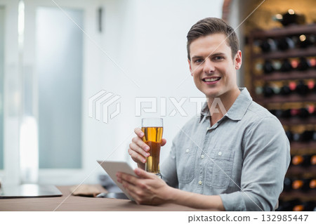Man wearing grey shirt sitting at bar counter holding beer glass, using tablet by wine rack Man wearing grey shirt sitting at bar counter holding beer glass, using tablet by wine rack 132985447