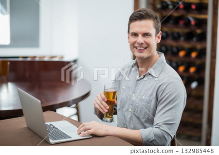 Man in late twenties typing on silver laptop holding beer glass at table in wine cellar Man in late twenties typing on silver laptop holding beer glass at table in wine cellar 132985448