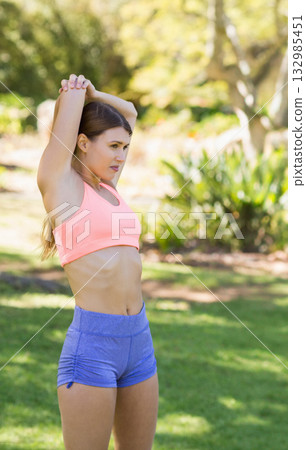 Female stretching arms overhead on sunlit park lawn wearing coral sports bra and blue shorts 132985451