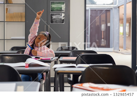 Female child student raising hand while writing in notebook at classroom desk with pen, copy space 132985457