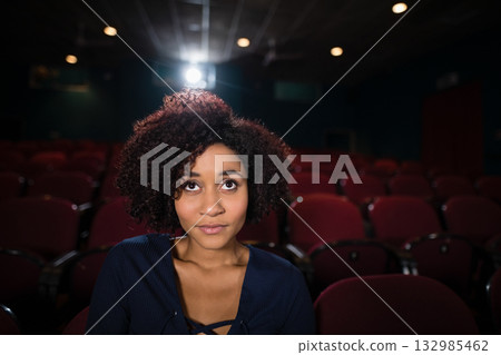 African American woman sitting facing screen in dim theater with red seats and projector light 132985462