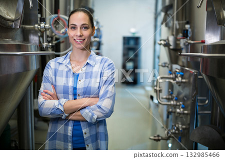 Female brewery technician standing in aisle between steel tanks with hoses, pipes, valves, smiling 132985466