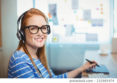 Woman in her twenties wearing headphones using stylus on drawing tablet at desk, copy space 132985474