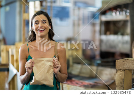 Female in her twenties smiling while holding brown paper bag inside rustic bakery with brick oven 132985478