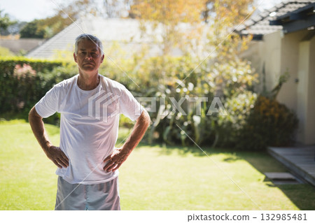 Senior man standing with hands on hips on backyard lawn near hedge under bright sunlight Senior man standing with hands on hips on backyard lawn near hedge under bright sunlight 132985481