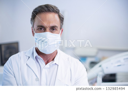 Male doctor standing in examination room wearing lab coat and surgical face mask Male doctor standing in examination room wearing lab coat and surgical face mask 132985494