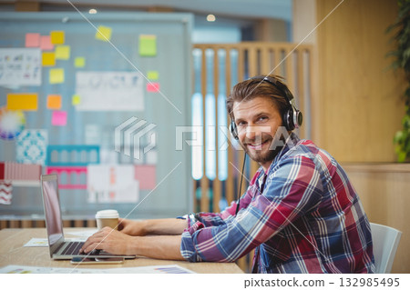 Man sitting at table in shared space wearing headphones using laptop, planning board, copy space Man sitting at table in shared space wearing headphones using laptop, planning board, copy space 132985495