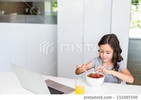 Girl sitting in home kitchen eating cereal holding spoon with laptop and orange juice, copy space Girl sitting in home kitchen eating cereal holding spoon with laptop and orange juice, copy space 132985500