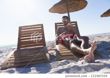 Man reclining on wooden lounge chair under straw parasol at beach with cocktail glass, copy space 132985504