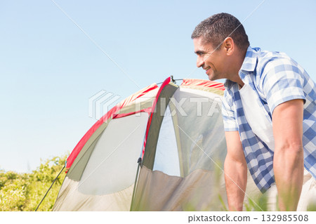 Middle-aged man leaning over red-trimmed dome tent entrance in grassy meadow and shrubs, copy space Middle-aged man leaning over red-trimmed dome tent entrance in grassy meadow and shrubs, copy space 132985508
