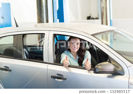 Female driver leaning out window of silver hatchback giving thumbs up near blue lifts at garage 132985517