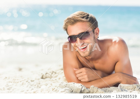 Shirtless man lying on white sand at beach by ocean wearing tinted sunglasses, smiling under sun Shirtless man lying on white sand at beach by ocean wearing tinted sunglasses, smiling under sun 132985519