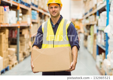 Male warehouse worker wearing hard hat and vest holding cardboard box in aisle among metal racks Male warehouse worker wearing hard hat and vest holding cardboard box in aisle among metal racks 132985521