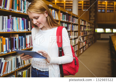 Female teenager standing in library aisle by bookshelves holding tablet and wearing red backpack 132985522