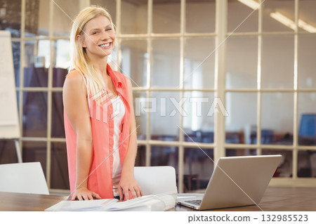 Woman smiling and leaning forward behind wooden conference table in office with laptop blueprints 132985523