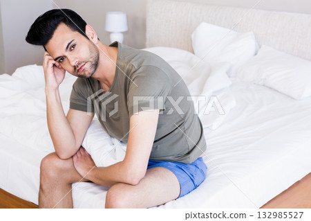 Mid adult man sitting on edge of unmade bed in bedroom with nightstand lamp holding head 132985527