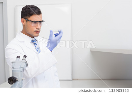 Male scientist wearing goggles and gloves examining sample container beside microscope in lab 132985533