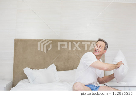 Man gripping pillow, looking right in bedroom on bed with white linens, headboard, copy space 132985535