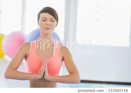 Woman wearing coral top standing with closed eyes pressing palms in yoga studio with exercise balls 132985536