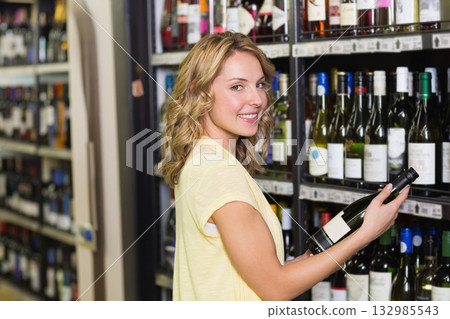 Woman holding green wine bottle, glancing back in liquor store aisle with metal shelves, copy space 132985543