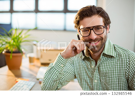 Bearded man wearing green shirt sitting at desk in office holding pen near keyboard, potted plant 132985546