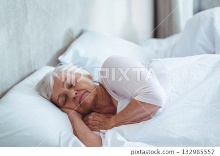 Senior woman lying on side in bed wearing sleep top and hugging white pillow against headboard Senior woman lying on side in bed wearing sleep top and hugging white pillow against headboard 132985557