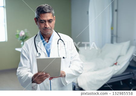 Male doctor wearing lab coat, stethoscope reviewing tablet in hospital room by bed, privacy curtain 132985558